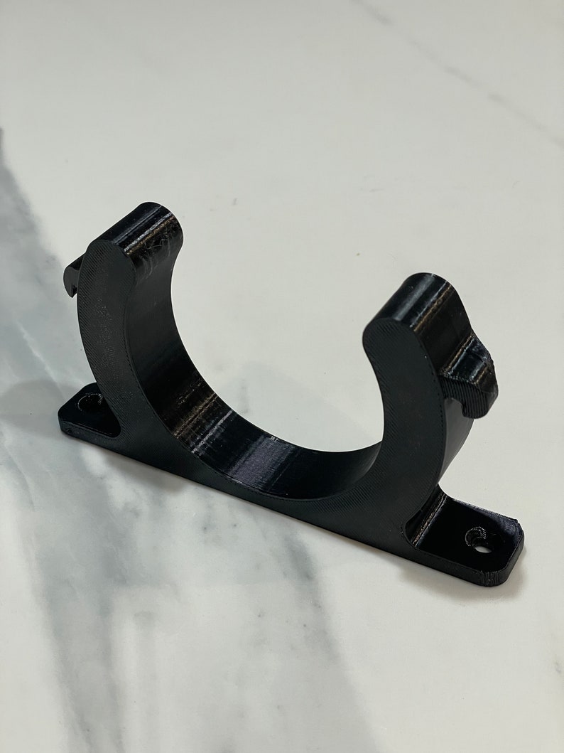May include: A black, 3D-printed bracket with a curved cradle and a rectangular base. The base has two holes for mounting. The bracket is designed to hold a cylindrical object. The material has visible layer lines.