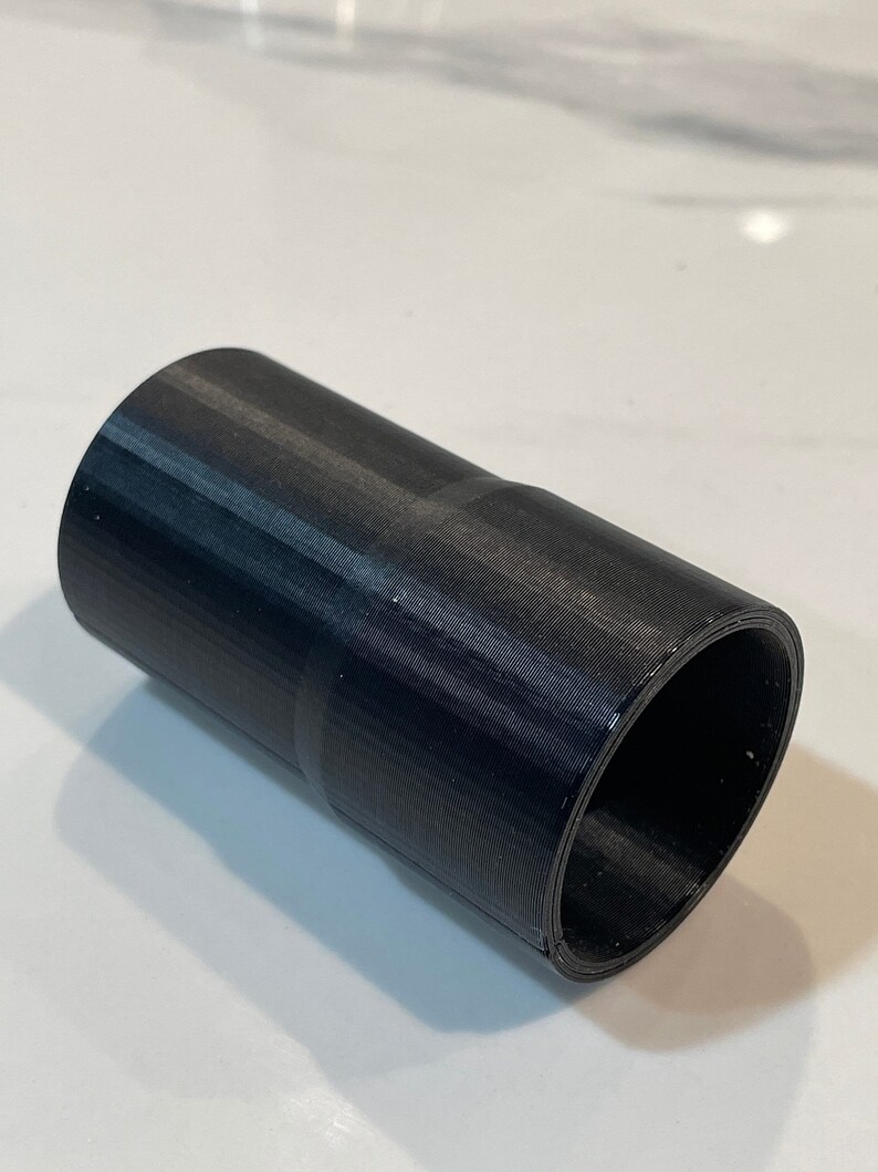 K&ouml;nnte beinhalten: Ein schwarzer zylindrischer Adapter mit strukturierter Oberfl&auml;che. Der Adapter hat an einem Ende eine breitere und am anderen Ende eine schmalere &Ouml;ffnung. Das Objekt besteht aus einem festen, dunklen Material.