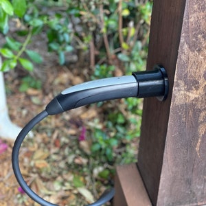 May include: An electric vehicle charging cable with a gray and black connector is plugged into a wooden post. The cable is black and curved, with green foliage in the background. The wooden post is brown.