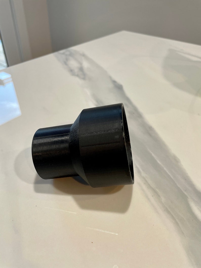 K&ouml;nnte beinhalten: Ein schwarzer Kunststoffadapter mit einem ausgestellten, zylindrischen Design. Der Adapter hat an einem Ende eine breitere &Ouml;ffnung und am anderen Ende eine schmalere &Ouml;ffnung, die durch einen konischen Abschnitt verbunden sind. Das Objekt wird wahrscheinlich zum Verbinden von Rohren oder Schl&auml;uchen unterschiedlicher Gr&ouml;&szlig;e verwendet.