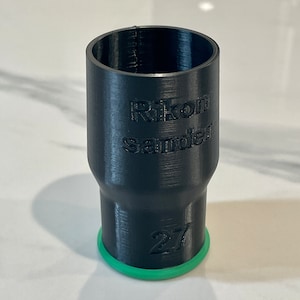 May include: A black cylindrical object with the text "Rikon Sander" and the number "27" printed on it. The base has a green ring. The object appears to be 3D-printed.