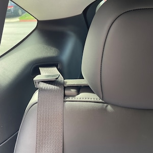 May include: Interior car shot featuring a gray seat with a seatbelt. The seatbelt is gray and extends from the top of the seat to a buckle. The headrest is gray and the interior is black.