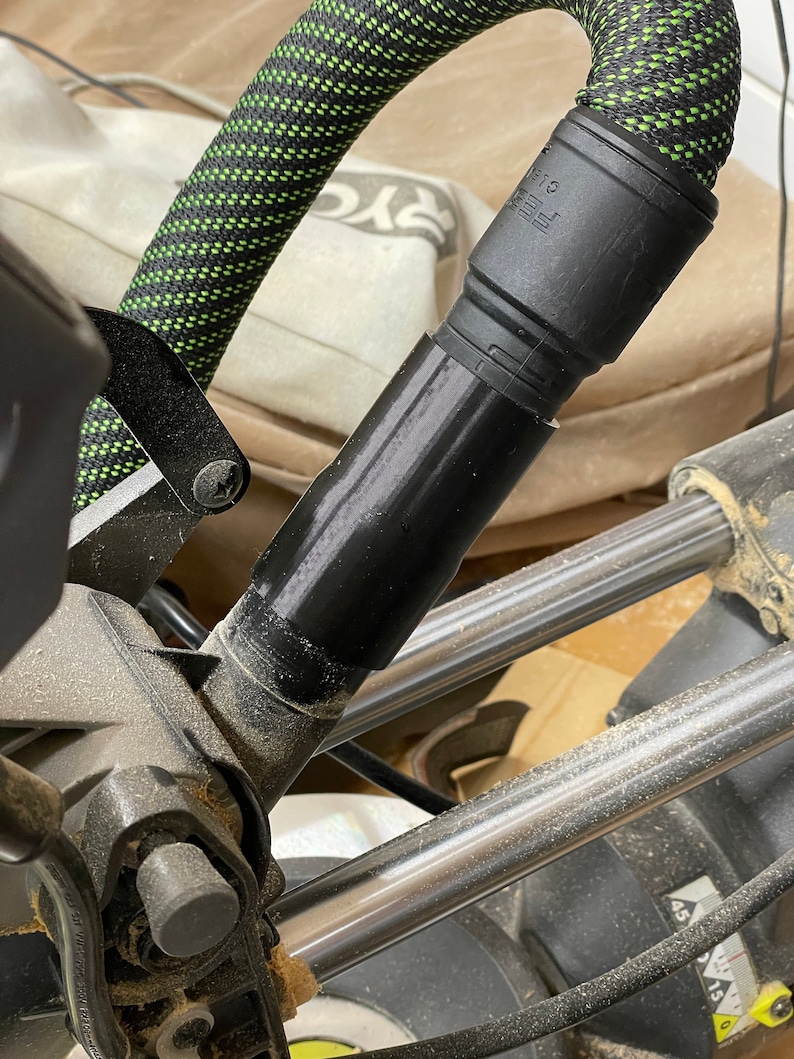K&ouml;nnte beinhalten: Nahaufnahme eines schwarzen und gr&uuml;nen Schlauchs, der mit einem schwarzen Adapter an einer Gehrungss&auml;ge befestigt ist. Der Schlauch hat ein schwarz-gr&uuml;nes Webmuster. Die S&auml;ge hat silberne Metallstangen und eine schwarze Basis. Detail eines Holzbearbeitungswerkzeugs.