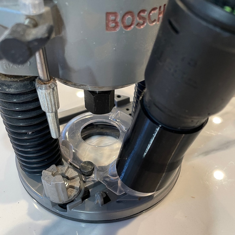 May include: A gray Bosch router with a clear plastic base and black accents. The word "BOSCH" is printed in red on the top of the tool. A black hose is attached to the side.