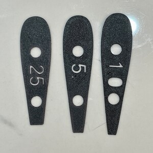 May include: Three black, teardrop-shaped measuring tools with white numbers. The tools have a hole at the top and bottom. The numbers are 25, 5, and 100.