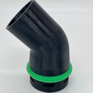 May include: A black, angled pipe fitting with a green circular seal. The fitting has a 45-degree bend and is likely 3D-printed. The green seal is a bright, contrasting color against the black plastic.