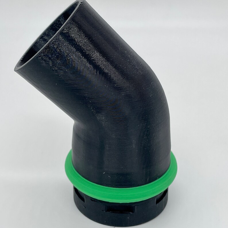 May include: A black, angled pipe fitting with a green circular seal. The fitting has a 45-degree bend and is likely 3D-printed. The green seal is a bright, contrasting color against the black plastic.