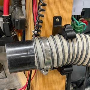 May include: A black plastic pipe is connected to a flexible, ribbed hose with a metal clamp. The hose is secured by a black bracket on a wooden post. Electrical wires and a coiled black cable are also visible.
