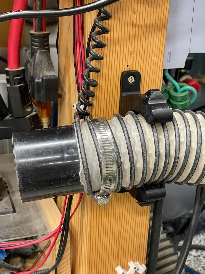 May include: A black plastic pipe is connected to a flexible, ribbed hose with a metal clamp. The hose is secured by a black bracket on a wooden post. Electrical wires and a coiled black cable are also visible.