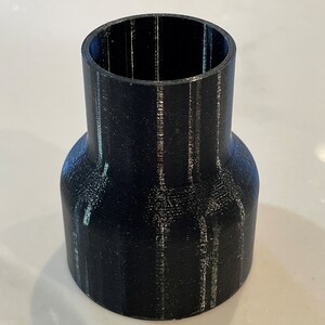 Puede incluir: Un adaptador cil&iacute;ndrico negro con una base ensanchada y una parte superior m&aacute;s estrecha. El adaptador parece estar impreso en 3D, con l&iacute;neas de capa visibles. El objeto est&aacute; dise&ntilde;ado para conectar tuber&iacute;as o accesorios de diferentes tama&ntilde;os.