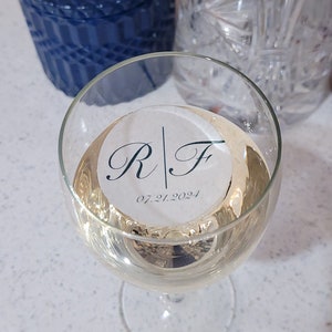 May include: A wine glass containing a pale liquid, possibly wine or champagne. A white circular sticker with the initials "R | F" and the date "07.21.2024" is inside the glass. Other glassware is visible in the background.