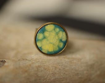 Puede incluir: Un anillo de latón con una incrustación redonda de esmalte azul y amarillo. La incrustación tiene un patrón abstracto en espiral.