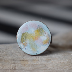 Puede incluir: Anillo redondo esmaltado con banda de cobre. El diseño esmaltado abstracto presenta colores como azul, amarillo, rosa y blanco. El esmalte tiene un aspecto texturizado y agrietado. Una pieza de joyería única.