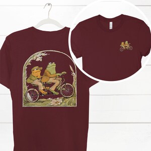 May include: A maroon t-shirt with a graphic of two frogs riding a tandem bicycle in a green, yellow, and brown landscape. The frogs are wearing hats and suits. The t-shirt has a crew neck and short sleeves.