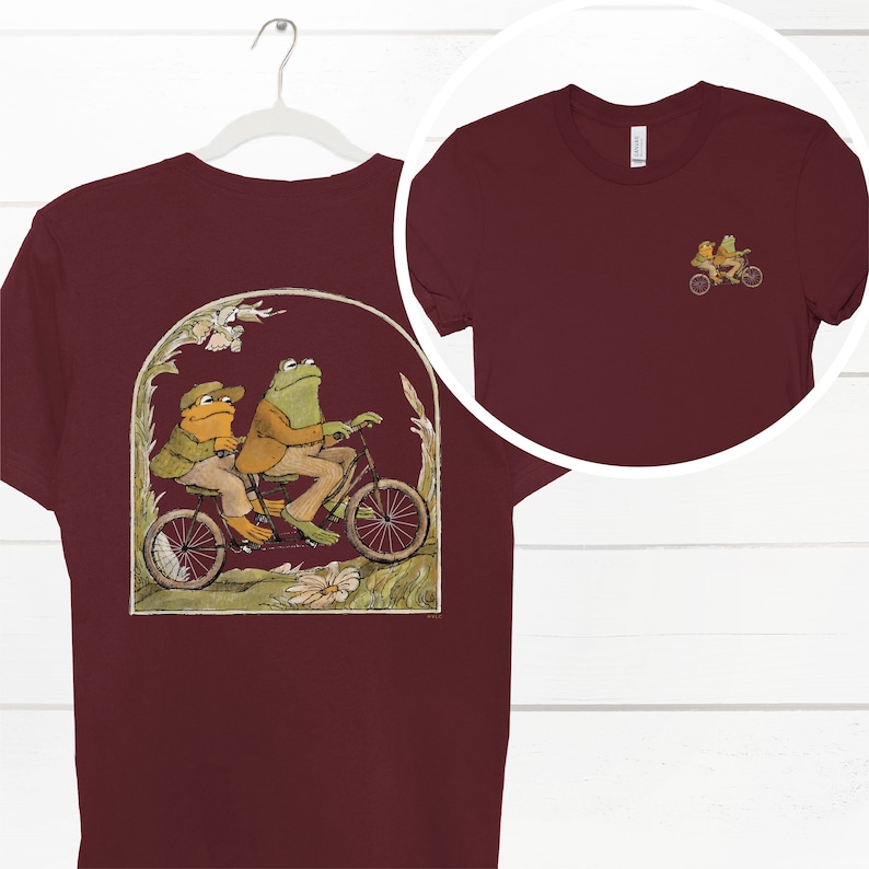 May include: A maroon t-shirt with a graphic of two frogs riding a tandem bicycle in a green, yellow, and brown landscape. The frogs are wearing hats and suits. The t-shirt has a crew neck and short sleeves.