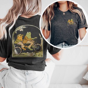 May include: A dark gray t-shirt with a graphic of two frogs riding a bicycle. The frogs are green and brown and are wearing clothes. The bicycle is red and has a basket on the front.