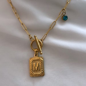 May include: A gold chain necklace with a toggle clasp and a rectangular pendant featuring the letter "M". A small blue gemstone dangles from the chain.