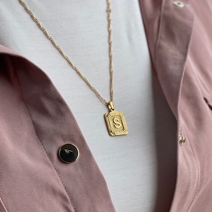 May include: A gold chain necklace with a gold pendant featuring the letter "S" in a square frame. The necklace is worn by a person wearing a white shirt and a pink jacket.