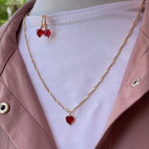 May include: A gold chain necklace with a red heart pendant and matching red heart earrings. The necklace and earrings are on a white shirt.