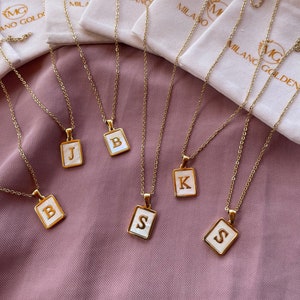 Op de afbeelding: Vijf gouden kettingen met witte rechthoekige hangers met de letters B, J, K en S. De kettingen worden gepresenteerd op een roze stoffen achtergrond met witte fluwelen zakjes.
