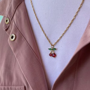 May include: A gold chain necklace with a red and green cherry pendant. The necklace is paired with matching cherry earrings.