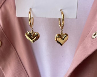 May include: Gold hoop earrings with a dangling heart charm. The heart charm is slightly puffed out and has a shiny finish.