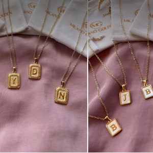 Op de afbeelding: Een verzameling gouden kettingen met initialen hangers. De hangers zijn rechthoekig en hebben een witte emaille achtergrond met gouden letters. De kettingen worden gepresenteerd op een roze stoffen achtergrond.