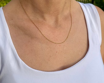 May include: A delicate gold-colored chain necklace resting on a person's chest. The necklace is thin and appears to be made of small, connected links. The person is wearing a white tank top.