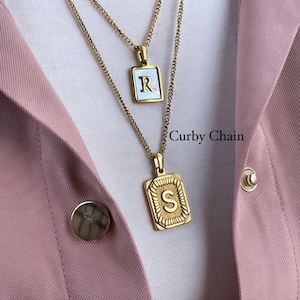 May include: Two gold necklaces with a curb chain style. The top necklace has a square pendant with the letter "R" in white on a gold background. The bottom necklace has a square pendant with the letter "S" in gold on a gold background. The pendant has a decorative border.