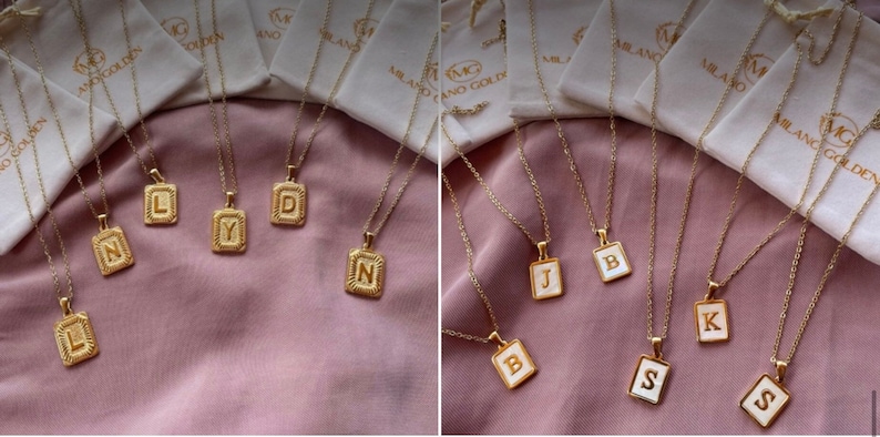 Op de afbeelding: Een verzameling gouden kettingen met initialen hangers. De hangers zijn rechthoekig en hebben een witte emaille achtergrond met gouden letters. De kettingen worden gepresenteerd op een roze stoffen achtergrond.