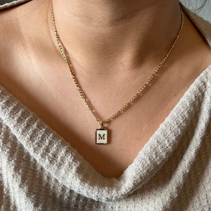 Op de afbeelding: Een gouden ketting met een witte vierkante hanger met de letter "M" in goud.