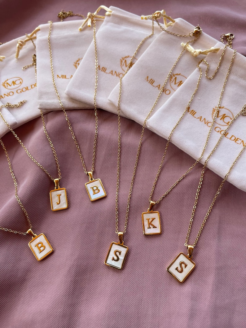 Op de afbeelding: Vijf gouden kettingen met witte rechthoekige hangers met de letters B, J, K en S. De kettingen worden gepresenteerd op een roze stoffen achtergrond met witte fluwelen zakjes.