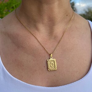 May include: A gold chain necklace with a square pendant featuring a detailed design. The pendant has a raised circle in the centre.