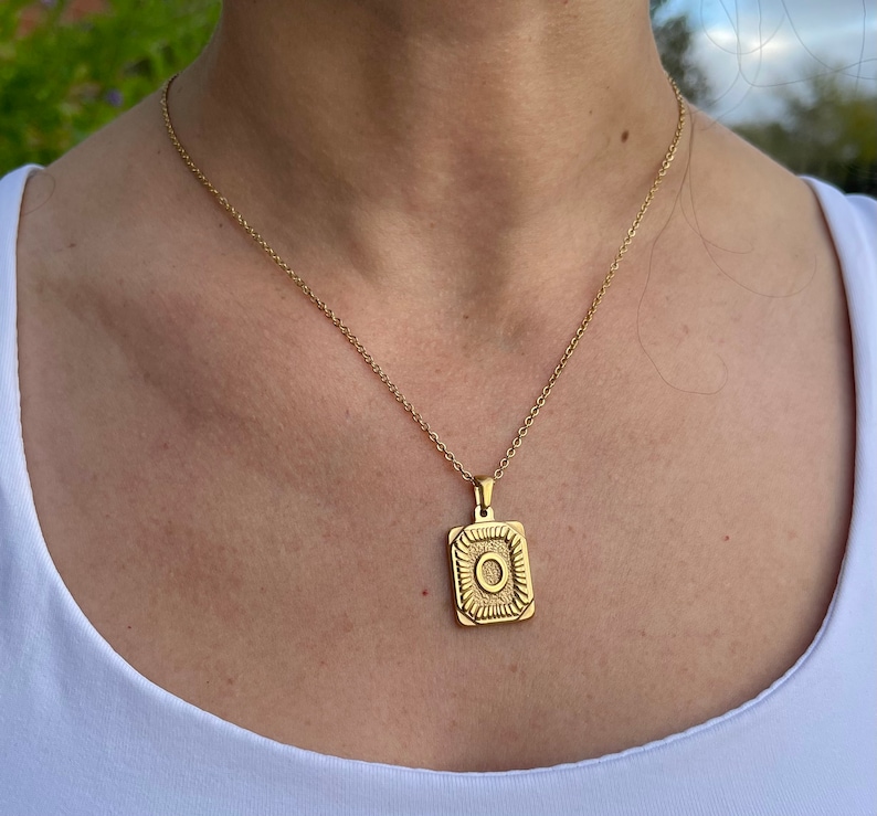 May include: A gold chain necklace with a square pendant featuring a detailed design. The pendant has a raised circle in the centre.