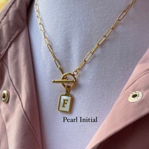 May include: A gold chain necklace with a toggle clasp and a square pendant featuring the letter "F" in white on a pearl-like background. The text "Pearl Initial" is visible below the pendant.