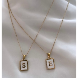 Op de afbeelding: Twee gouden kettingen met witte emaille hangers. De hangers zijn vierkant en hebben de letters "S" en "E" in goud.