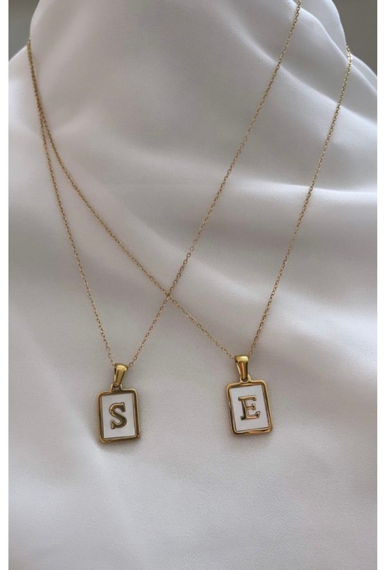 Op de afbeelding: Twee gouden kettingen met witte emaille hangers. De hangers zijn vierkant en hebben de letters "S" en "E" in goud.