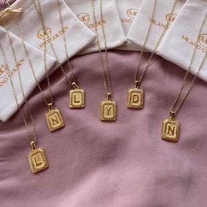Op de afbeelding: Zes gouden kettingen met vierkante hangers met de letters L, Y, D, N en N. De kettingen liggen op een paarse stoffen ondergrond.