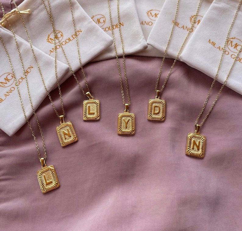 Op de afbeelding: Zes gouden kettingen met vierkante hangers met de letters L, Y, D, N en N. De kettingen liggen op een paarse stoffen ondergrond.