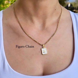 Op de afbeelding: Een gouden Figaro-ketting met een klein vierkant hanger met een witte achtergrond en de letter "B" in goud.