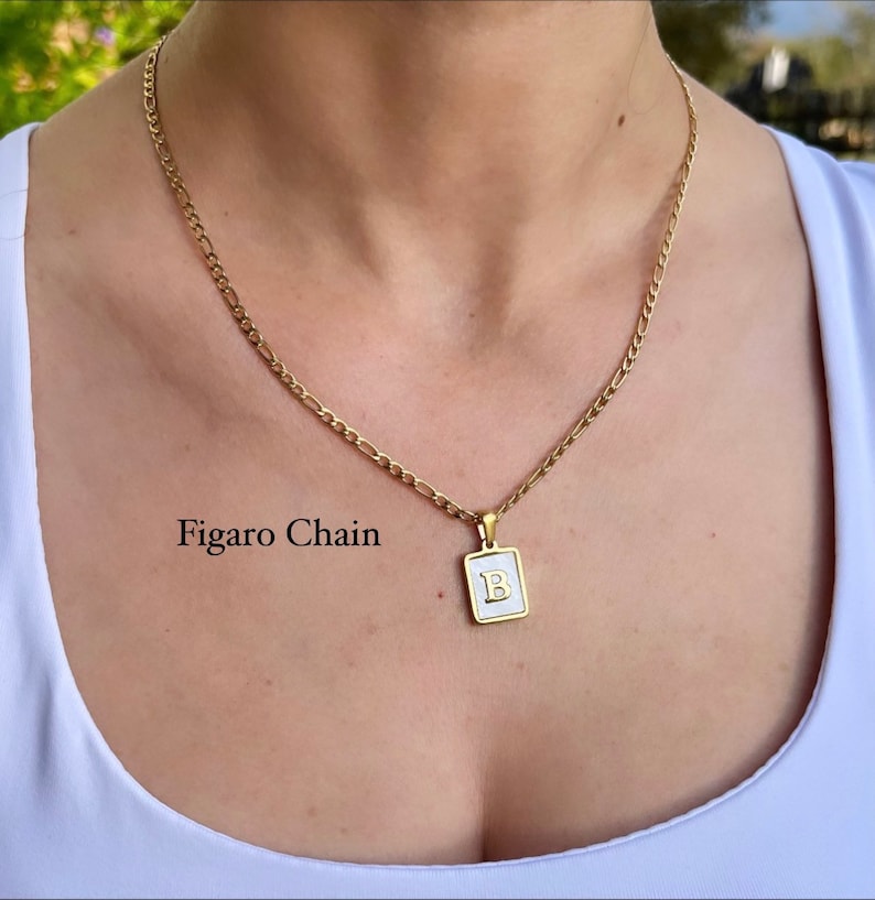 Op de afbeelding: Een gouden Figaro-ketting met een klein vierkant hanger met een witte achtergrond en de letter "B" in goud.