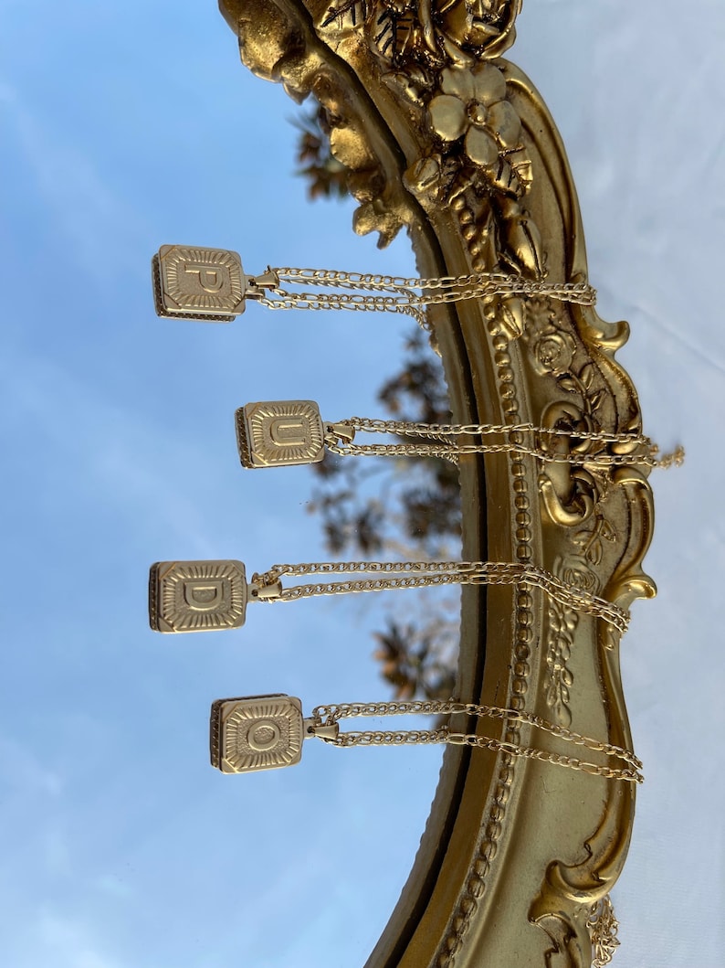 May include: Four gold necklaces with square pendants featuring the letters P, U, D, and O. The necklaces are hanging from a gold-framed mirror.