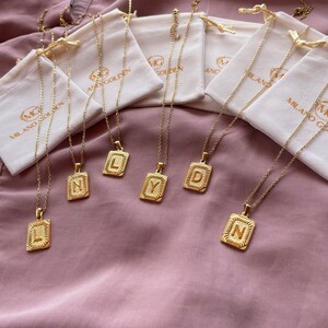 May include: Six gold necklaces with initial pendants in the letters L, Y, D, N, and two other letters. The necklaces are displayed on a pink fabric background and each necklace is in a small white pouch with the text "Milano Golden" printed on it.