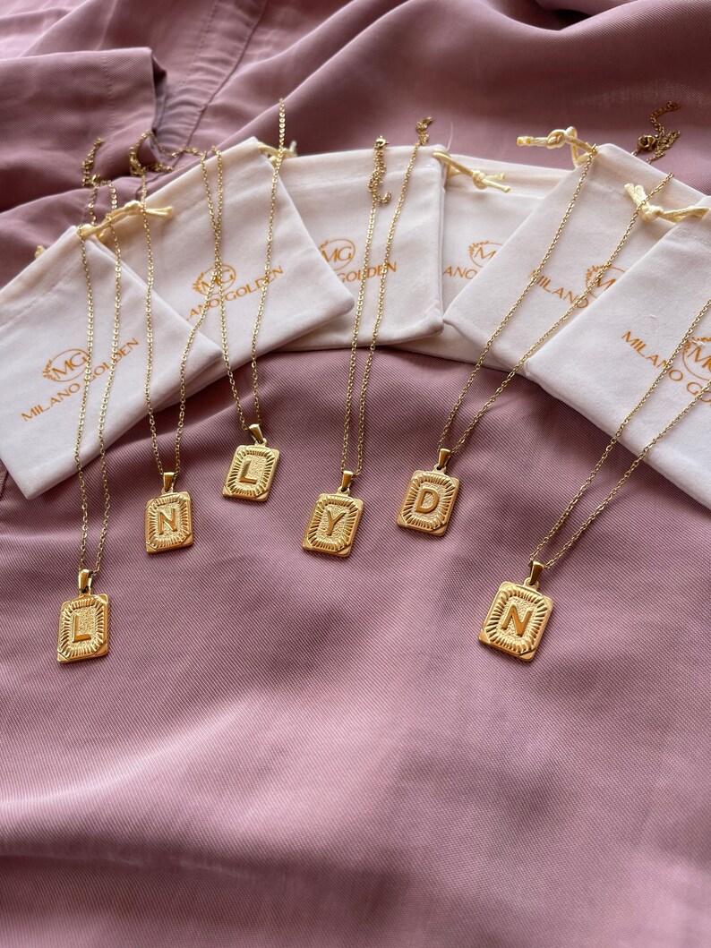 May include: Six gold necklaces with initial pendants in the letters L, Y, D, N, and two other letters. The necklaces are displayed on a pink fabric background and each necklace is in a small white pouch with the text "Milano Golden" printed on it.