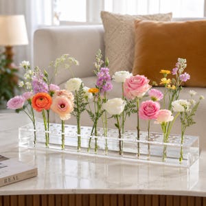 Peut inclure: Un long vase rectangulaire en acrylique transparent doté de multiples ouvertures circulaires individuelles sur la surface supérieure. Le vase contient une variété de fleurs fraîches, notamment des roses roses, des renoncules orange et des mufliers violets.