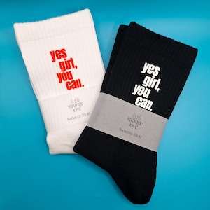 Könnte beinhalten: Zwei Paar Crew-Socken, eine weiße und eine schwarze, mit dem Aufdruck "yes girl, you can" in kontrastierenden Farben. Die Socken sind mit einem Papierband mit Markennamen und Größe umwickelt.