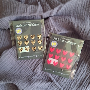 Könnte beinhalten: Zwei Packungen Bügelflicken. Eine Packung enthält Leopardenmuster-Herz-Patches, die andere rosa Herz-Patches. Die Packungen sind mit "Patch zum Aufbügeln" mit "DIY" in einem Kreis beschriftet.