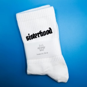 Könnte beinhalten: Weiße Socken mit dem Wort "sisterhood" in schwarzer Schrift. Die Socken haben ein Etikett mit "little strange love" und "Socken Gr. 39-41". Auf blauem Hintergrund.