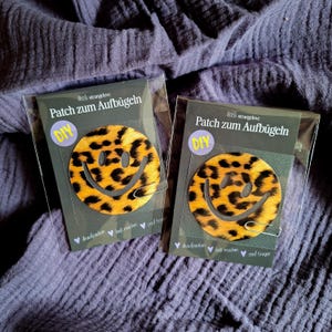 Könnte beinhalten: Zwei Aufbügler mit einem Smiley-Gesicht im Leopardenmuster in Gelb und Schwarz. Jeder Aufbügler ist in einer transparenten Plastikhülle mit dem Text "Patch zum Aufbügeln" und "DIY" verpackt. Auf einem lila Stoffhintergrund.