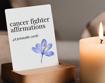 Może przedstawiać: Biała karta z tekstem "cancer fighter affirmations" i "48 printable cards". W prawym dolnym rogu karty znajduje się niebieski kwiat z złotymi akcentami. Karta stoi na drewnianym stojaku. Na tle znajduje się biała świeca.